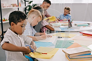 selective focus of teacher and interracial preschoolers cutting colorful papers with scissors