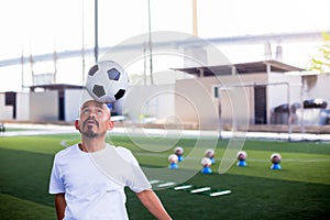 Selective focus to ball on head of football coach to demo for football player with blurry marker cones and soccer equipment