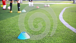 Selective focus to blue marker cone is soccer training equipment on green artificial turf with blurry kid players training backgro