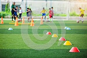 Selective focus to red and yellow marker cones are soccer training equipment on green artificial turf with blurry kid players trai