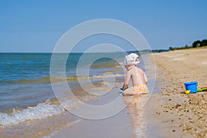 Selective focus on toddler girl sitting in the water on the sand on the beach. The child plays with toys. Back view. On
