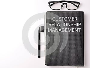 Selective focus top view text CUSTOMER RELATIONSHIP MANAGEMENT on black book with pen and eye glasses isolated on white background