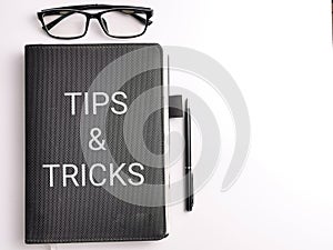 Selective focus top view text TIPS AND TRICKS on black book with pen and eye glasses isolated on white background.