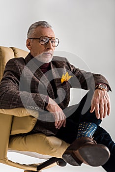 Selective focus of trendy mature man in glasses sitting in armchair with crossed legs isolated