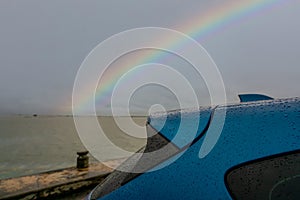 Selective focus on trunk of blue luxury SUV car with raindrops on rainbow and stormy sky background. Car parked at outdoor car