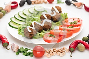 Selective focus of vegetables arranged in round frame around falafel with sauce on plate on white background.