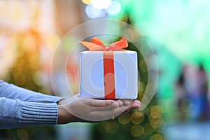 Selective focus of Woman hands holding white gift box with orange ribbon on bokeh abstract background for Christmas and New