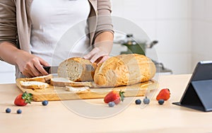 Selective focus woman or wife hand holding knife, cutting bread in cozy home kitchen in morning, preparing healthy meal for