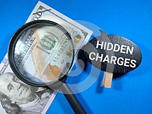 Selective focus.Word HIDDEN CHARGES on black board with magnifying glass and dollar banknote on blue background.Business concept.