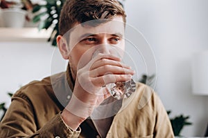 Selective focus of young caucasian man drinking fresh water from glass, sit on couch in room at home. Health care, proper