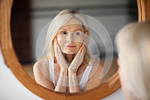 Self-care concept. Beautiful senior woman looking at mirror, touching soft skin on face and enjoying her reflection