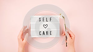 Self-care word on lightbox and flower narcissus on pink background flat lay. Take care of yourself