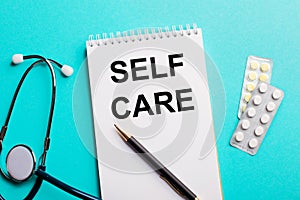 SELF CARE written in a white notepad near a stethoscope, pens and pills on a light blue background. Medical concept