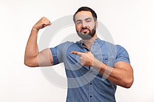 Self confident egotistical bearded man in blue shirt pointing finger at his arm biceps, proud and satisfied with muscular build, photo