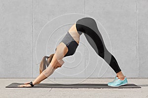 Self determined fitness woman does sport exercises on mat in gym, stands on hands, dressed in tanktop and leggings, poses against