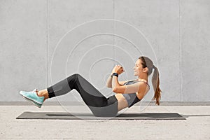 Self determined sporty woman with pony tail, dressed in leggings, top, sneakers, smartwatch makes on press, wants to have muscular