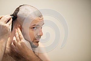 Self haircut, a man cuts his head with a clipper at home in front of a mirror