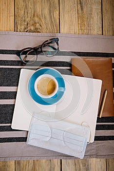 Self-isolation working at home flat lay: leather book, open blank pages of paper notebook, a pencil, glasses, medic mask and a cup