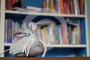 Self-made stuffed toy mouse in front of a colorful chaotic bookshelf . Reading mouse with blue eyes , mustache hair and hangdog