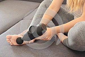 Self-massage of women's legs with a perfecting gun at home. Athletic woman applying therapeutic percussive massage gun