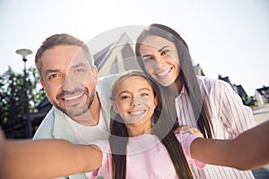 Self-portrait of attractive cheerful family mommy daddy embracing youth daughter having fun in cottage town outdoor