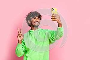 Self portrait of young blogging arabian guy holding smartphone shows v sign into cadre wear hoodie isolated on pink