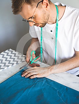 A self-taught seamster in white t-shirt and glasses works with fabric, pattern and chalk. A tailor cuts fabric in a home studio.