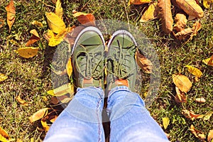 Selfie legs close-up in blue jeans and green sneakers standing on green grass with yellow leaves in autumn park
