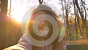 Selfie-photo of curly-haired caucasian girl watching earnestly into camera in sunny autumnal park.