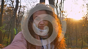 Selfie-photo of curly-haired caucasian girl watching smilingly into camera and winking at it in sunny autumnal park. photo