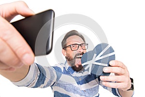Selfie portrait of attractive, crazy bearded man in jeans shirt shooting selfie with heart over white background.
