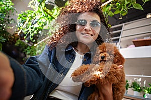 Selfie portrait brunette woman with curly hair holding small brown poodle dog and smiling indoors. Real happy female