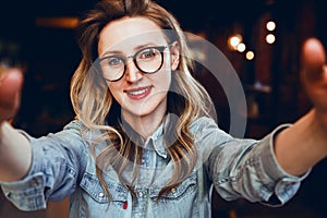 Selfie portrait of young smiling woman sitting in cafe. Hipster girl in trendy glasses takes a selfie in coffee shop.