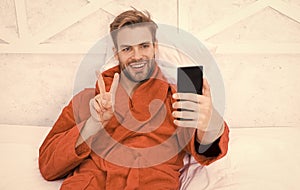 Selfie star. Happy man taking selfie with smartphone in bed. Handsome guy smiling with V hand gesture to selfie camera