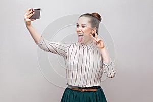 Selfie time! Portrait of happy foolish joyful attractive blogger woman wearing in striped shirt standing, winking and showing