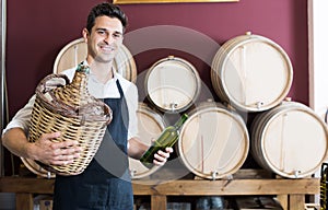 Seller in apron holding big wicker bottle with wine in store
