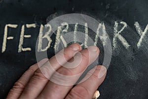 The semi-erased word FEBRUARY on the black chalkboard. An adult man`s left hand removes the handwritten word with his fingers.
