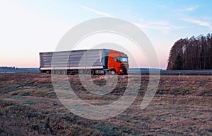A semitrailer tractor with a tilt semitrailer transports cargo against the backdrop of an evening sunny sunset in summer