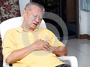 Senile asian men sitting on a chair at living room with heart attacks.Both old man`s hands on breast because of hard breathing photo