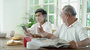 Senior 70s elderly Asian couple has breakfast. Man reading newspaper while woman using mobile phone.