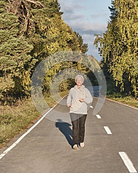 Senior active woman with gray hair running along forest paved road on sunny autumn day.