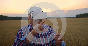 Senior adult farmer in a virtual reality helmet in a field of grain crops. In the sunset light, an elderly man in a