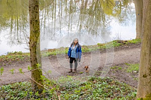 Senior adult female hiker standing next to her brown short-haired dachshund against water with mirror reflection
