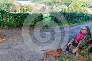 Senior adult woman sitting on wooden bench with her dachshund looking at camera