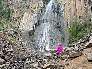 Senior Adult Woman Wearing a Fuchsia Jacket Sitting on a Boulder at the Base of a Waterfall