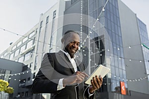 A senior African American businessman is talking to his team via video call. Standing near an office center in a suit