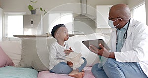 Senior african american male doctor in face mask using tablet talking to boy patient, slow motion