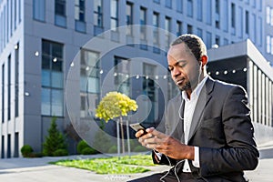 A senior African-American man, a businessman in a suit sits outside an office center wearing headphones and listening to