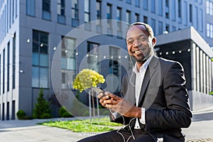 A senior African-American man, a businessman in a suit sits outside an office center wearing headphones and listening to