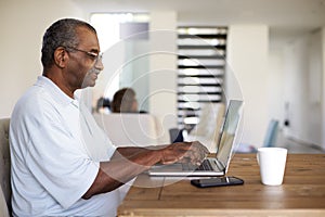 Senior African American  man sitting at the table using a laptop computer at home, side view